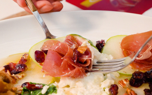 Carpaccio de Pera y Jamón Serrano: láminas finas de pera con jamón serrano, arúgula, arándanos secos, nueces y queso sobre un plato blanco.