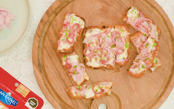 Cheesy Sticks con Jamón Real: pan tostado cubierto con queso derretido y trozos de jamón sobre una tabla de madera.