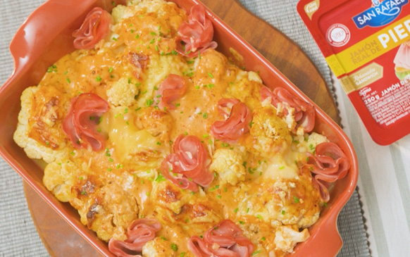 Coliflor gratinada con jamón: coliflor horneada con queso derretido, decorada con flores de jamón de pierna en una fuente de cerámica roja.